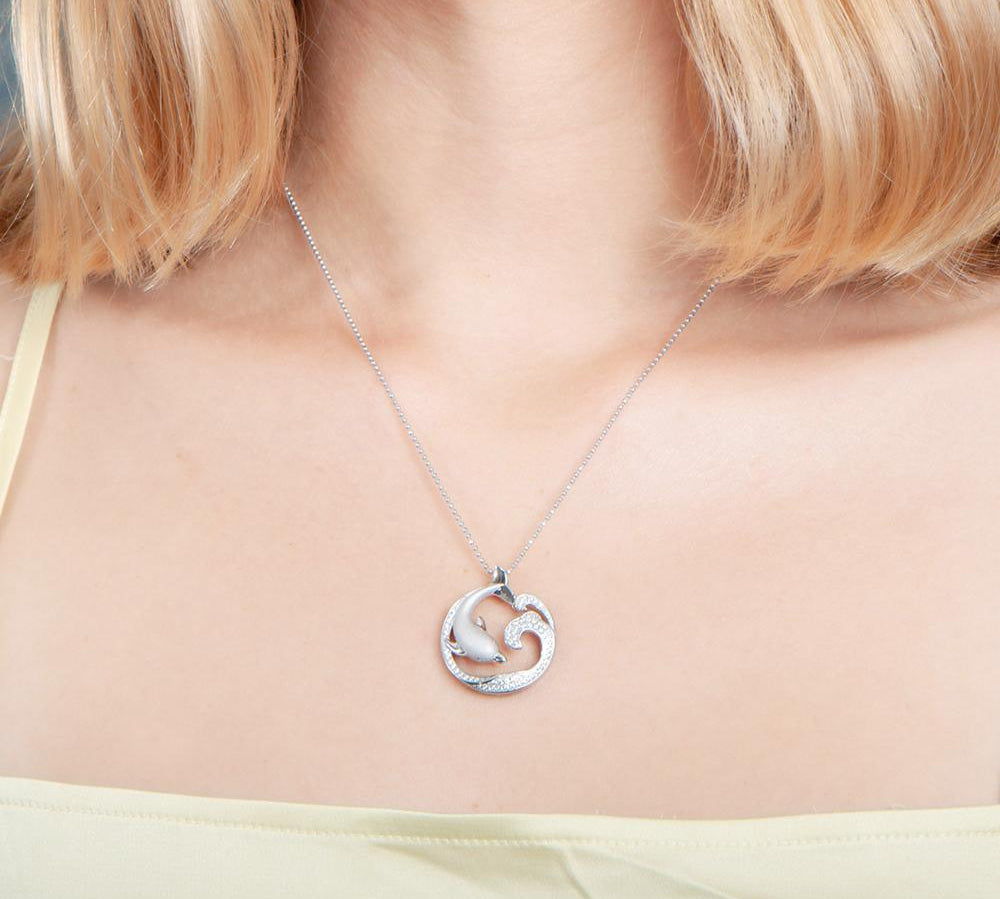 The picture shows a model wearing a 925 sterling silver, white gold vermeil, dolphin and wave pendant with topaz.