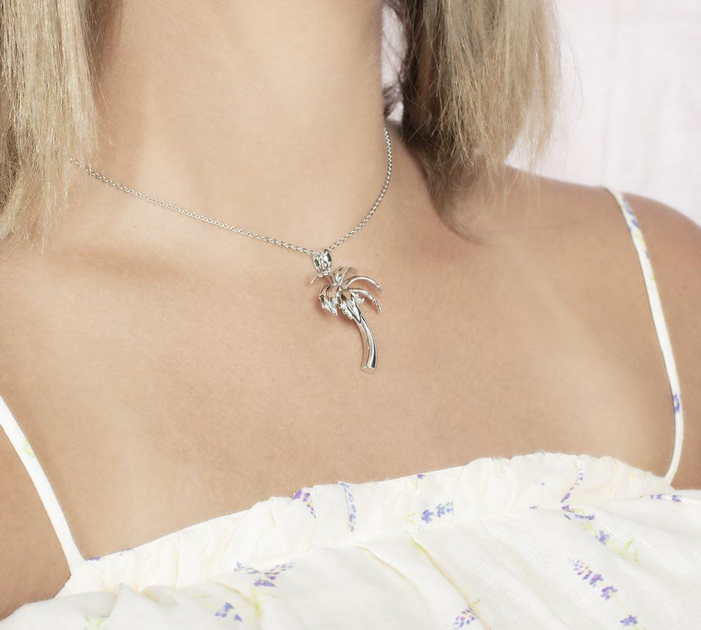 In this photo there is a model with blonde hair and white shirt with purple flowers turned to the right, wearing a sterling silver palm tree pendant with topaz gemstones.