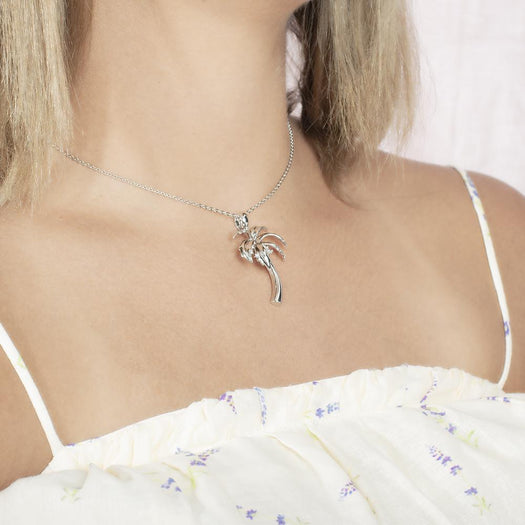 In this photo there is a model with blonde hair and white shirt with purple flowers turned to the right, wearing a sterling silver palm tree pendant with topaz gemstones.
