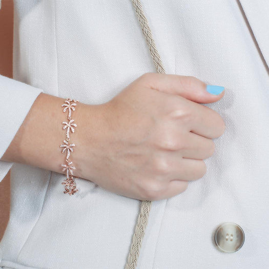In this photo there is a close-up of a model with a white coat and blue nail polish holding a beige purse strap, wearing a rose gold palm tree bracelet with topaz gemstones.