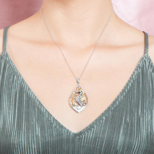 The picture shows a model wearing a two-tone white and yellow gold sea horse mandorla pendant with aquamarine and topaz.
