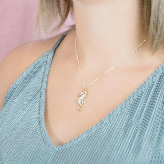 In this photo there is a model turned to the left with blonde hair and a teal shirt, wearing a yellow gold seahorse pendant with topaz gemstones.