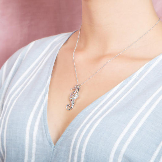 In this photo there is a model turned to the left with a light blue and white striped shirt, wearing a sterling silver seahorse pendant.