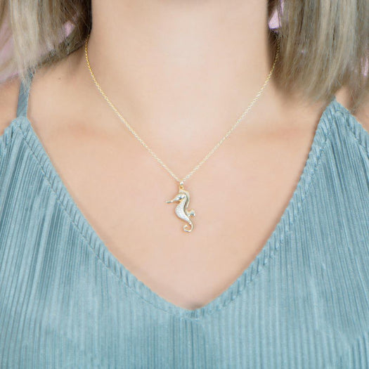 In this photo there is a model with blonde hair and a teal shirt, wearing a yellow gold seahorse pendant with topaz gemstones.
